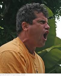 A white man looking angry and yelling, standing outside in yellow shirt.