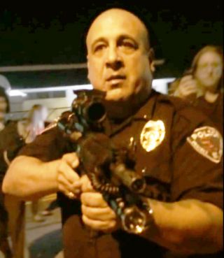 Large policeman holding large rifle pointing it at civilians.