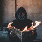 A masked, hooded figure holds up a burning newspaper